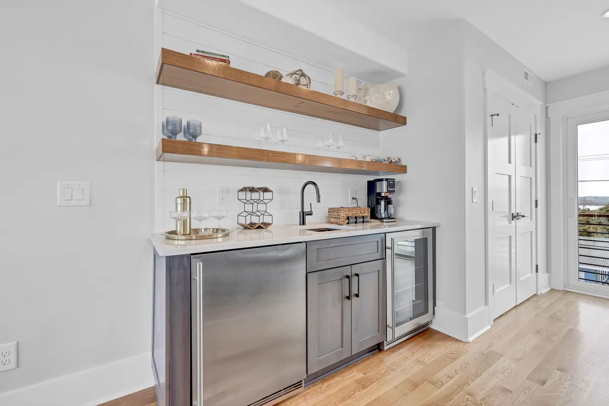 A stylish wet bar area featuring a bar sink, open shelving, coffee maker, and decorative accents. The compact but elegant setup is ideal for preparing drinks or morning coffee, supporting entertaining and social moments.