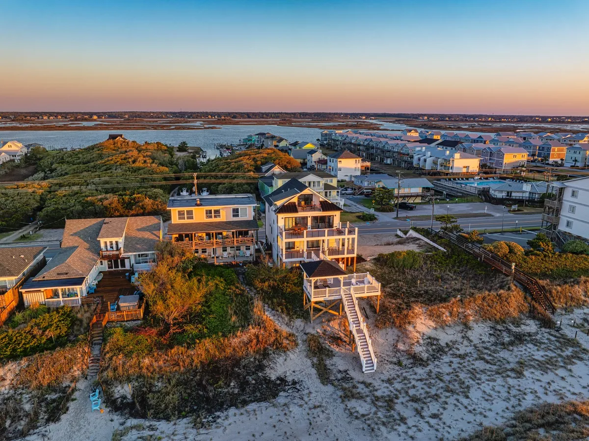 A beachfront house stands at the edge of soft sand, with steps leading directly from the deck to the inviting beach below. The surrounding landscape features gentle dunes and a glimpse of the neighborhood beyond, emphasizing direct, private access to the shoreline.