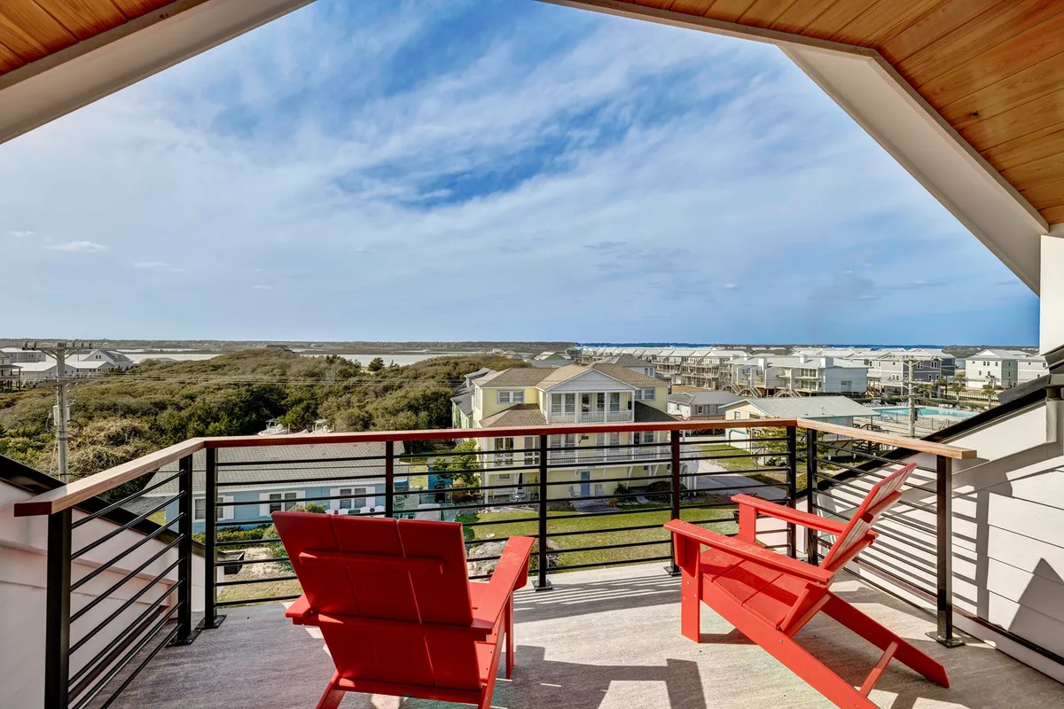 This image features a set of red Adirondack chairs positioned on a deck, overlooking the ocean and a landscape of dunes and neighboring houses. The vantage point suggests a private space to relax while enjoying sweeping coastal views, emphasizing the connection between the home's exterior living areas and the surrounding seascape.
