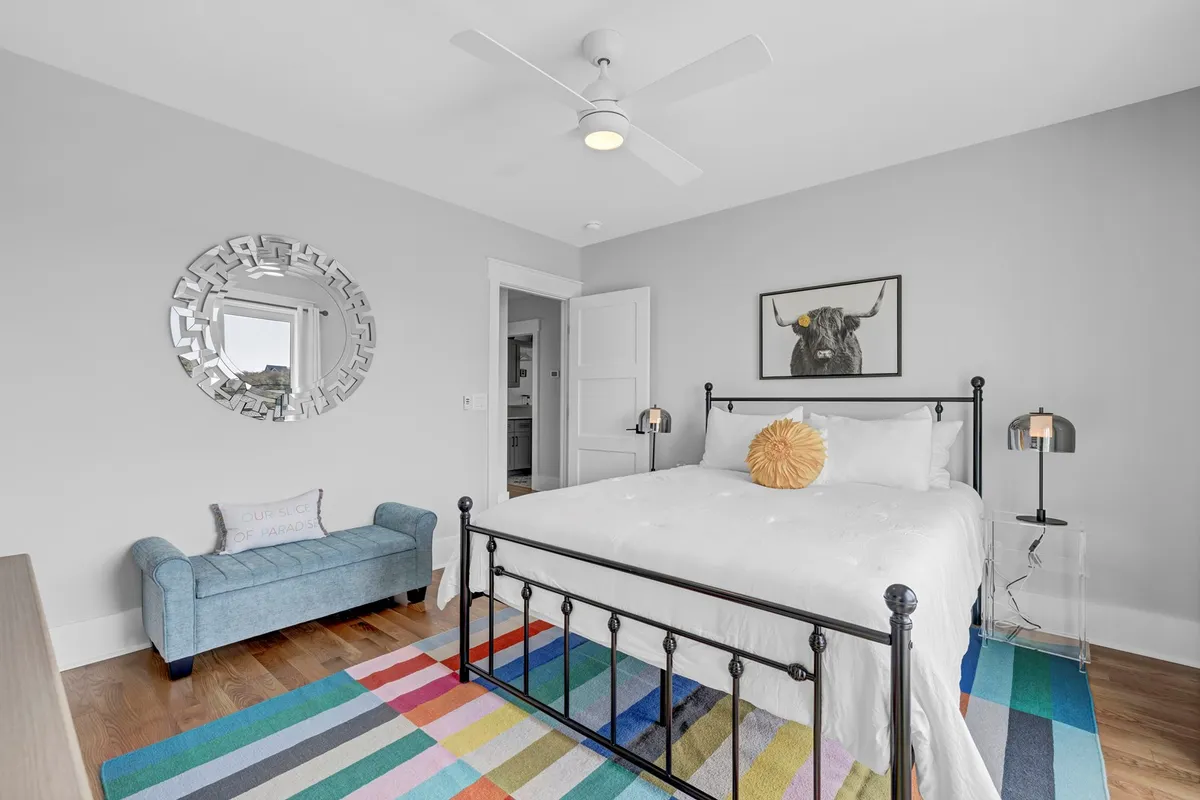 A vibrant queen bedroom adorned with colorful accents, a decorative pillow on the bed, a blue upholstered bench, and unique wall art. The room is designed for relaxation, with ample natural light and a welcoming vibe.