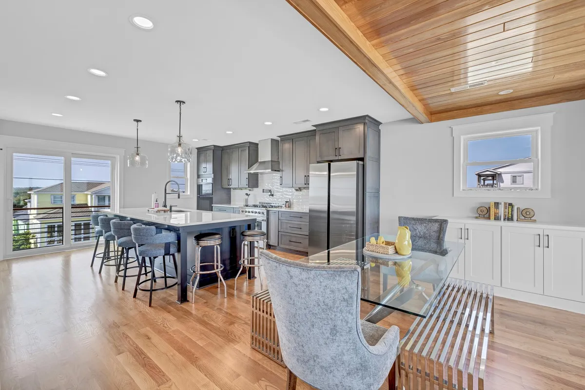 A modern, well-equipped kitchen with island bar seating, sleek appliances, a dining table, wooden ceiling, and expansive windows. The open layout seamlessly connects the kitchen to dining and living areas, emphasizing both function and style for entertaining and family meals.