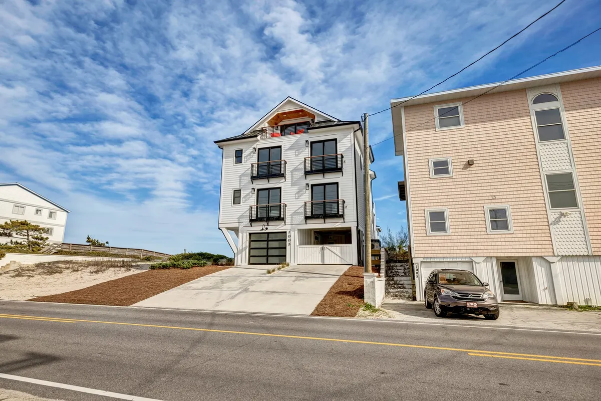 A cheerful exterior view of the modern vacation home, showcasing multiple balconies with railings, a welcoming driveway, and open spaces ideal for family gatherings. The architecture suggests generous, multi-level accommodations perfect for group stays.