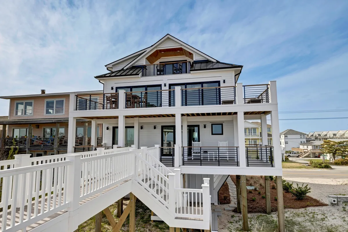 The hero image showcases a spacious, newly-built single-family beach house with expansive decks and balconies lined with white railings. Large windows flood the home with natural light. The outdoor spaces invite guests to dine al fresco, relax in the sun, and gather together while overlooking the serene surroundings.