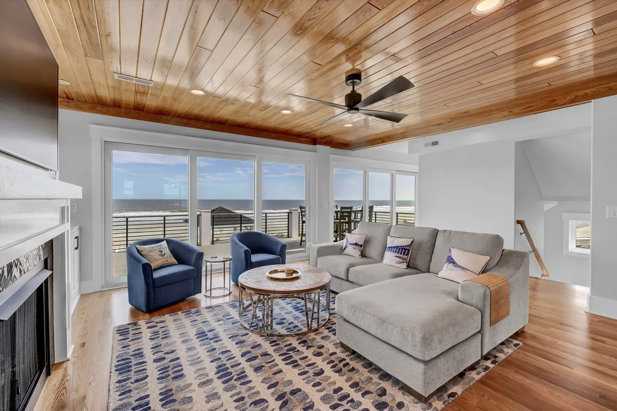 A beautifully designed open-concept living room featuring a comfortable gray sectional sofa, large windows offering sweeping ocean views, a textured area rug, wooden ceiling, and a decorative coffee table. The space is bathed in natural light and invites relaxation or group gatherings.